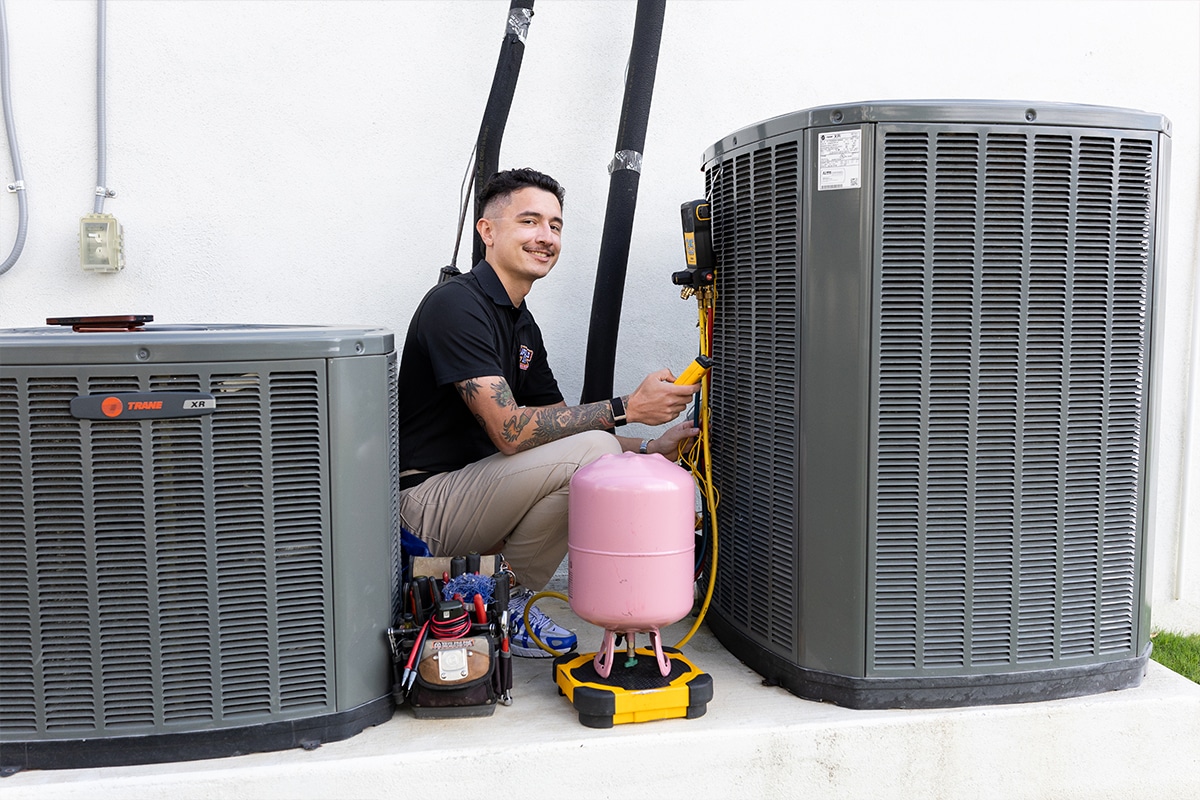 Smiling Apollo Air technician connecting a Fieldpiece manifold gauge to a Trane XR outdoor AC unit with a refrigerant tank and tools nearby