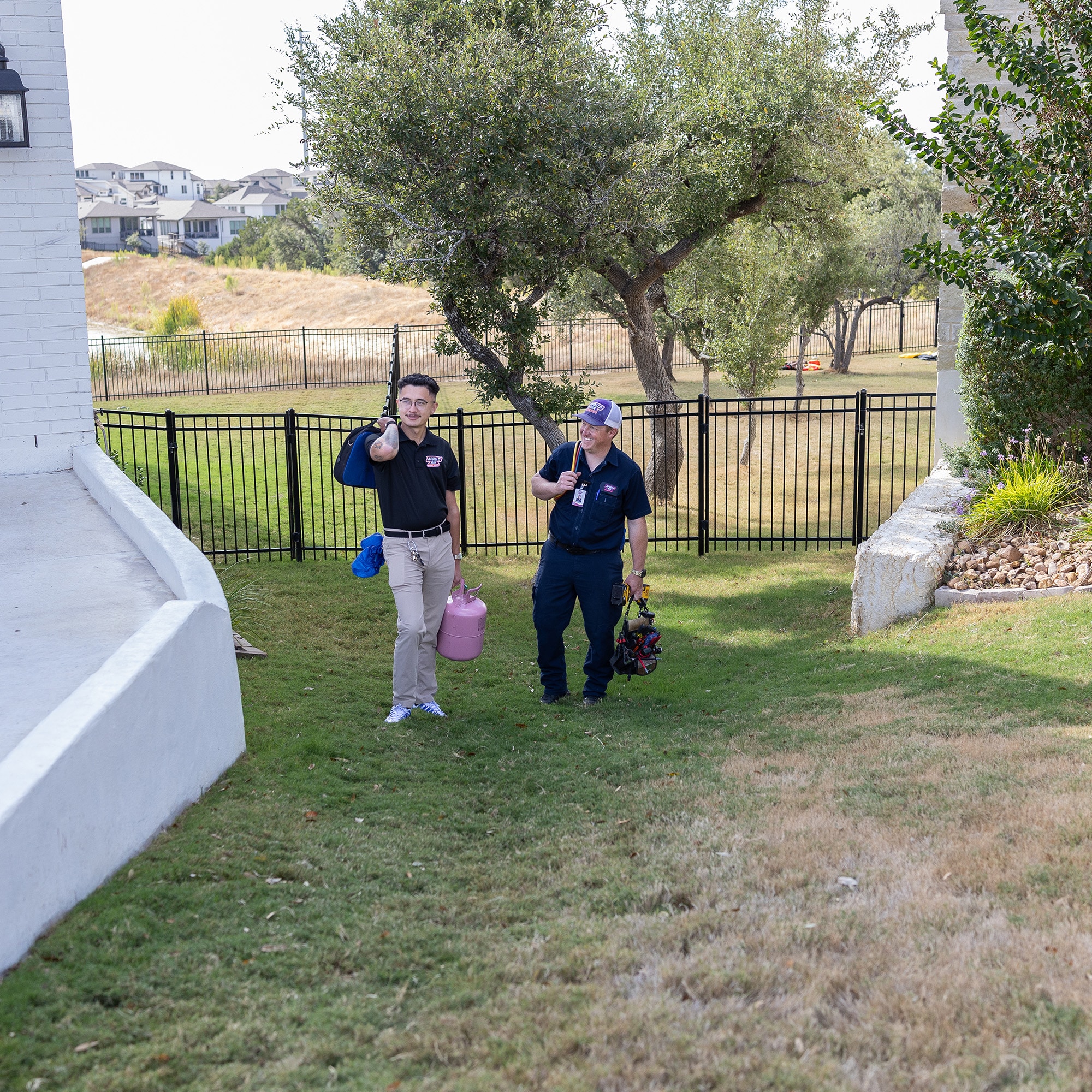 Two Apollo Air HVAC technicians walking through a residential backyard carrying a refrigerant tank and tool bag for an AC service call