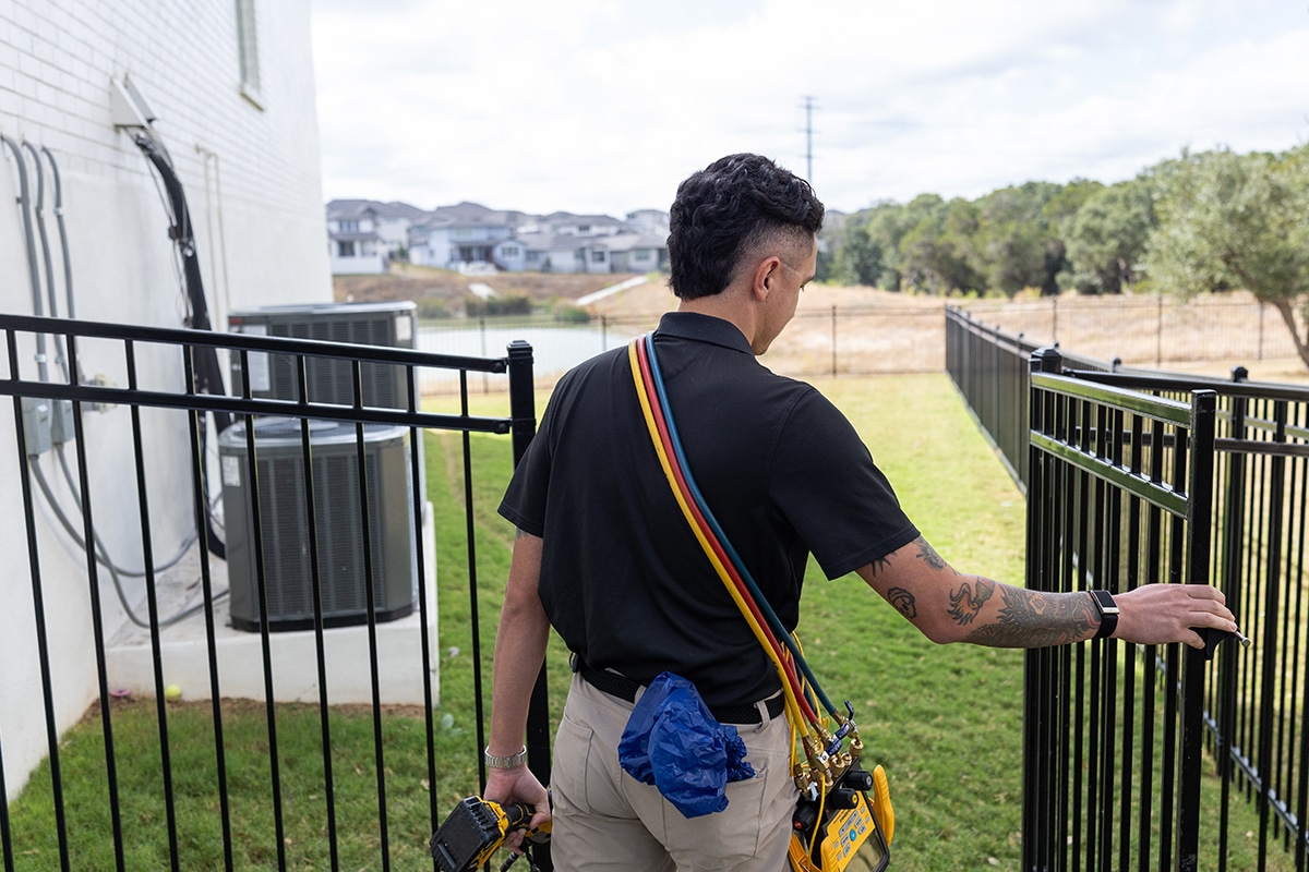 Apollo Air HVAC technician carrying refrigerant manifold gauge and drill entering backyard gate to service outdoor AC unit