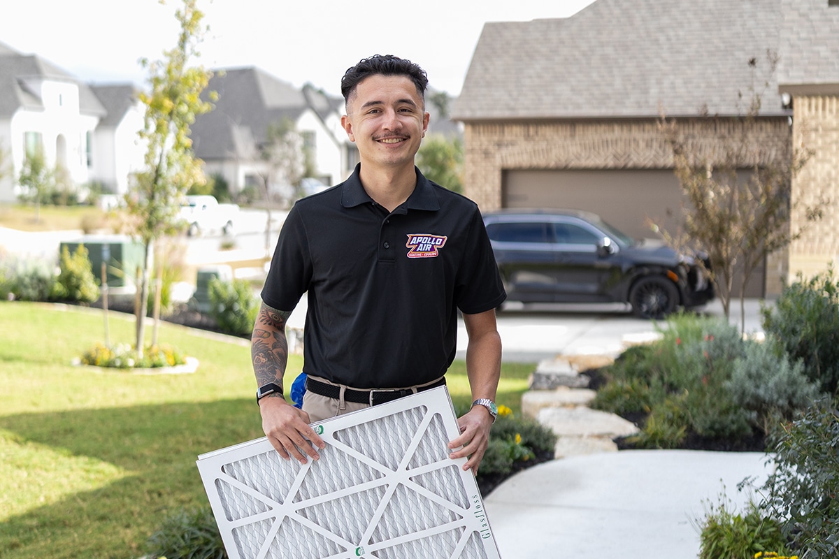 Smiling Apollo Air Heating and Cooling technician holding a Glasfloss air filter in front of a residential home ready for an HVAC maintenance service