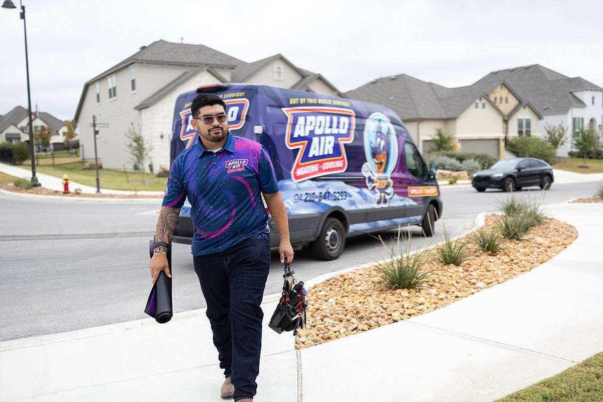 Apollo Air Heating and Cooling technician in branded uniform walking toward a residential home carrying tools with company van in background